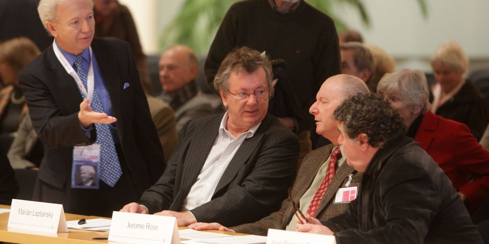 Several men are sitting at a table while a man in a suit is standing and speaking. Name tags on the table read 'Marian Lapšanský' and 'Jerome Rose'.