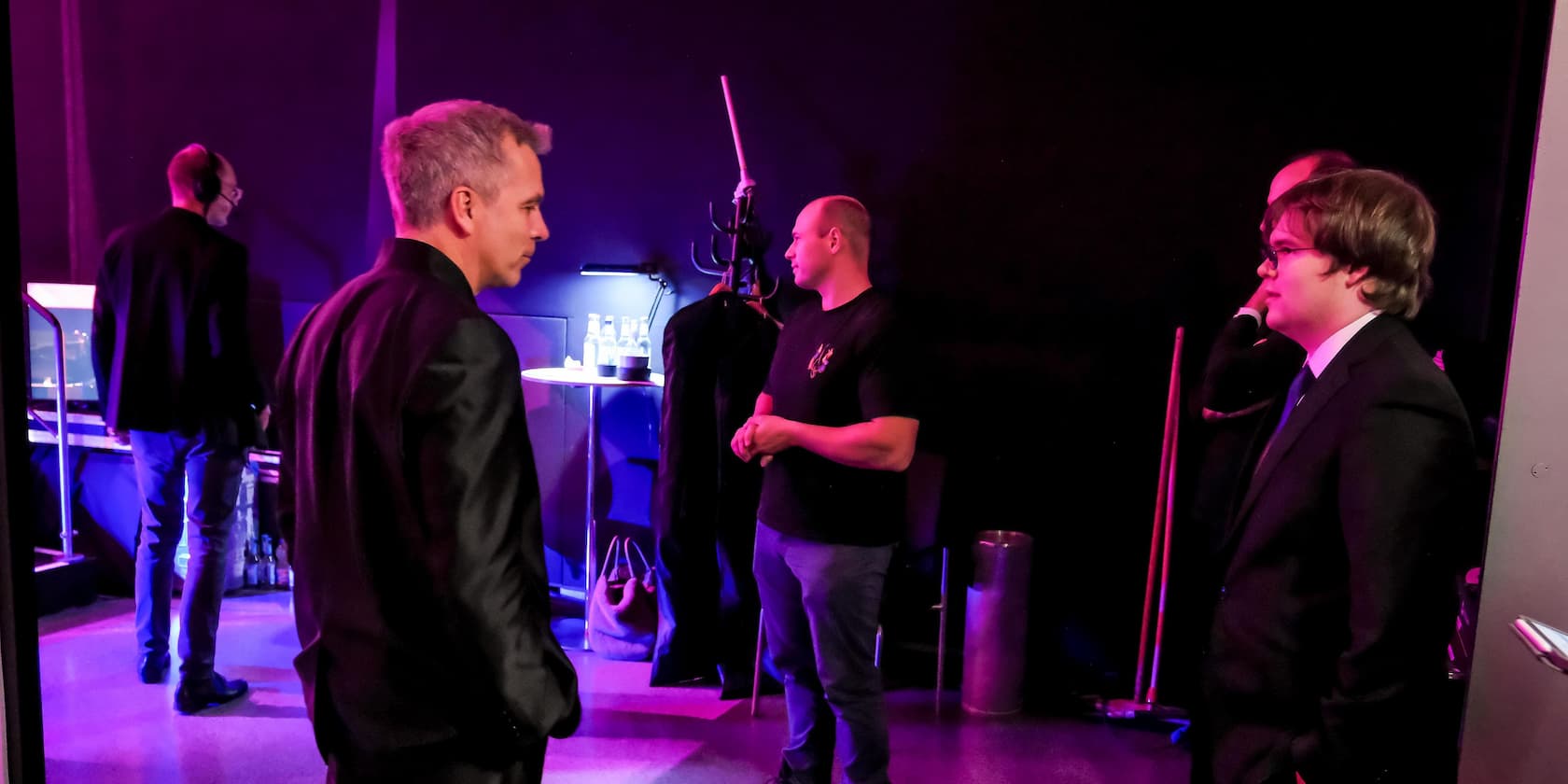 Several men in formal attire are talking backstage at an event.