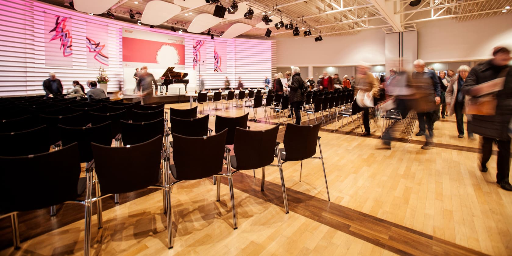 People entering a modern event hall with chairs and a grand piano on the stage.