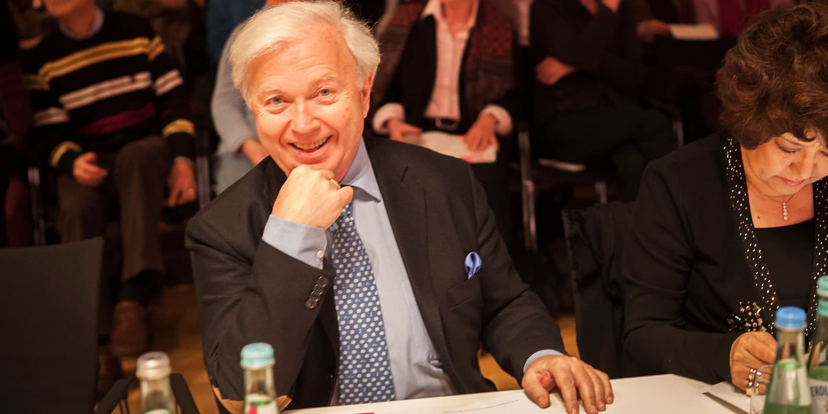 An elderly man in a suit smiles at the camera while sitting at a table in a crowded hall.