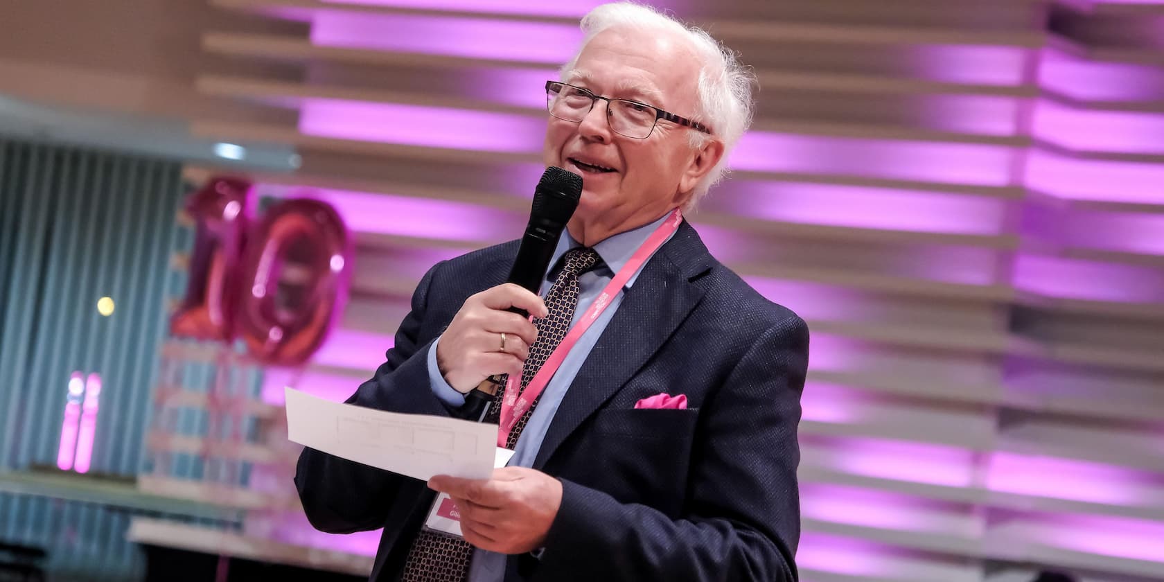 An elderly man holds a microphone and a note. In the background, balloons with the number 10 and purple lighting are visible.