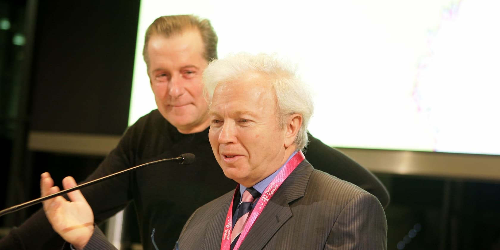 Two elderly men standing next to each other, speaking into a microphone at an event.