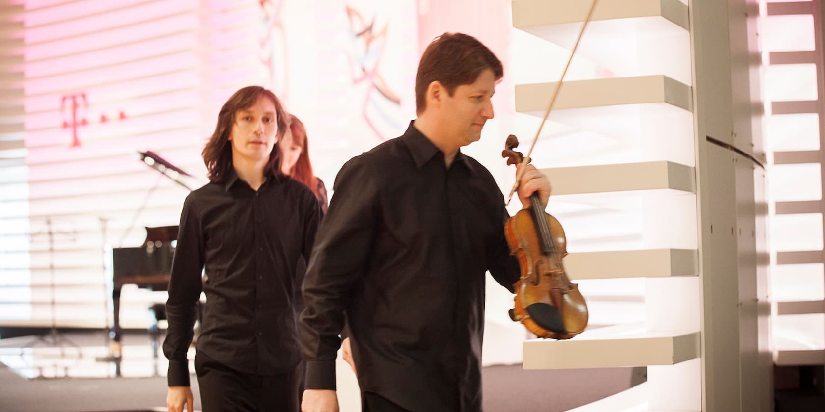 Two musicians in black shirts on a stage. One of them is carrying a violin.