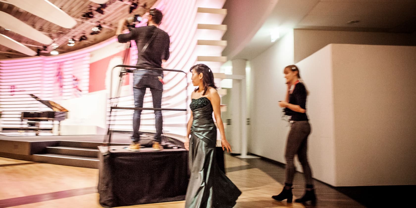 A cameraman films on an elevated platform while a woman in a green dress and a woman with a headset walk by.