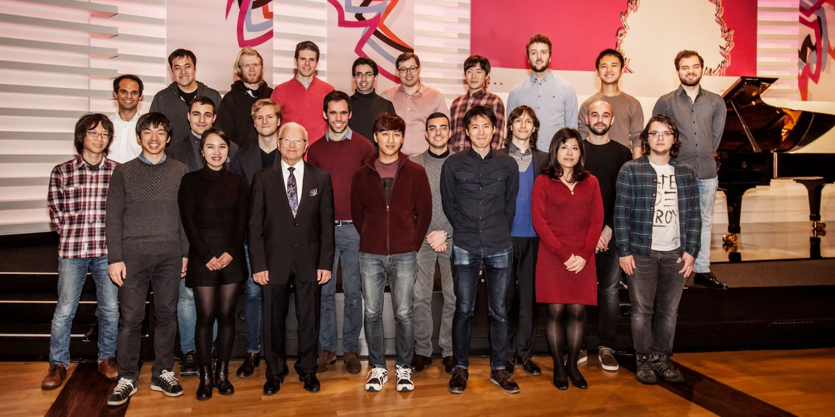 Group of 22 people posing for a photo on a stage; a grand piano in the background.