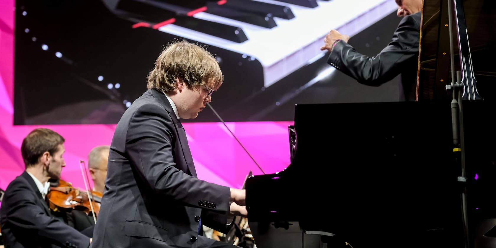 A man plays the piano at a concert with an orchestra in the background. A large screen displays a close-up of piano keys.