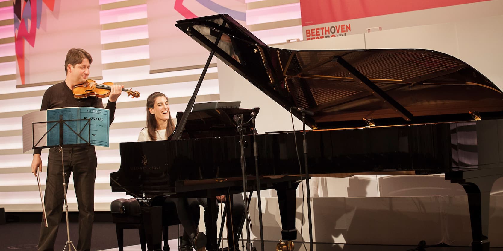 A violinist and a pianist performing on stage. Music stand displays Beethoven sonatas for piano and violin.