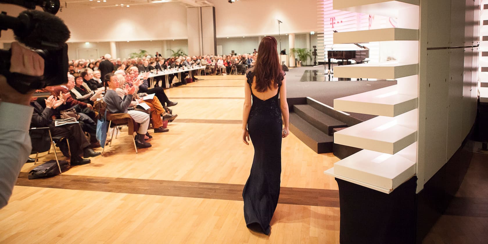 A woman in an elegant dress walks towards a stage as the audience applauds.