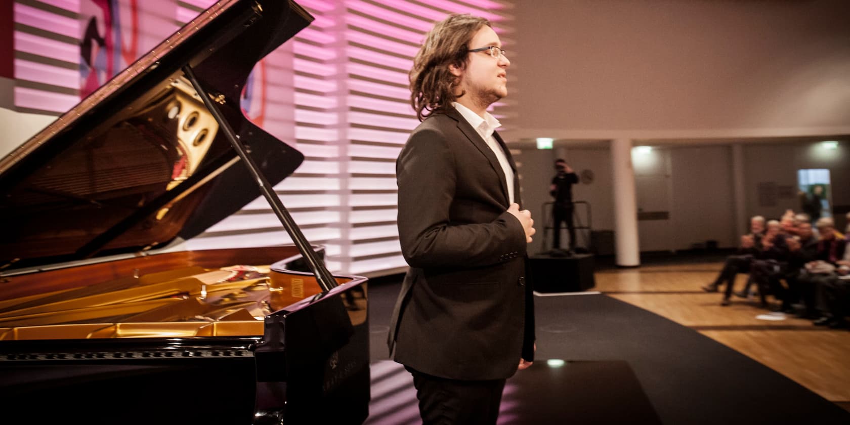 A man in a black suit stands next to a grand piano in a concert hall.