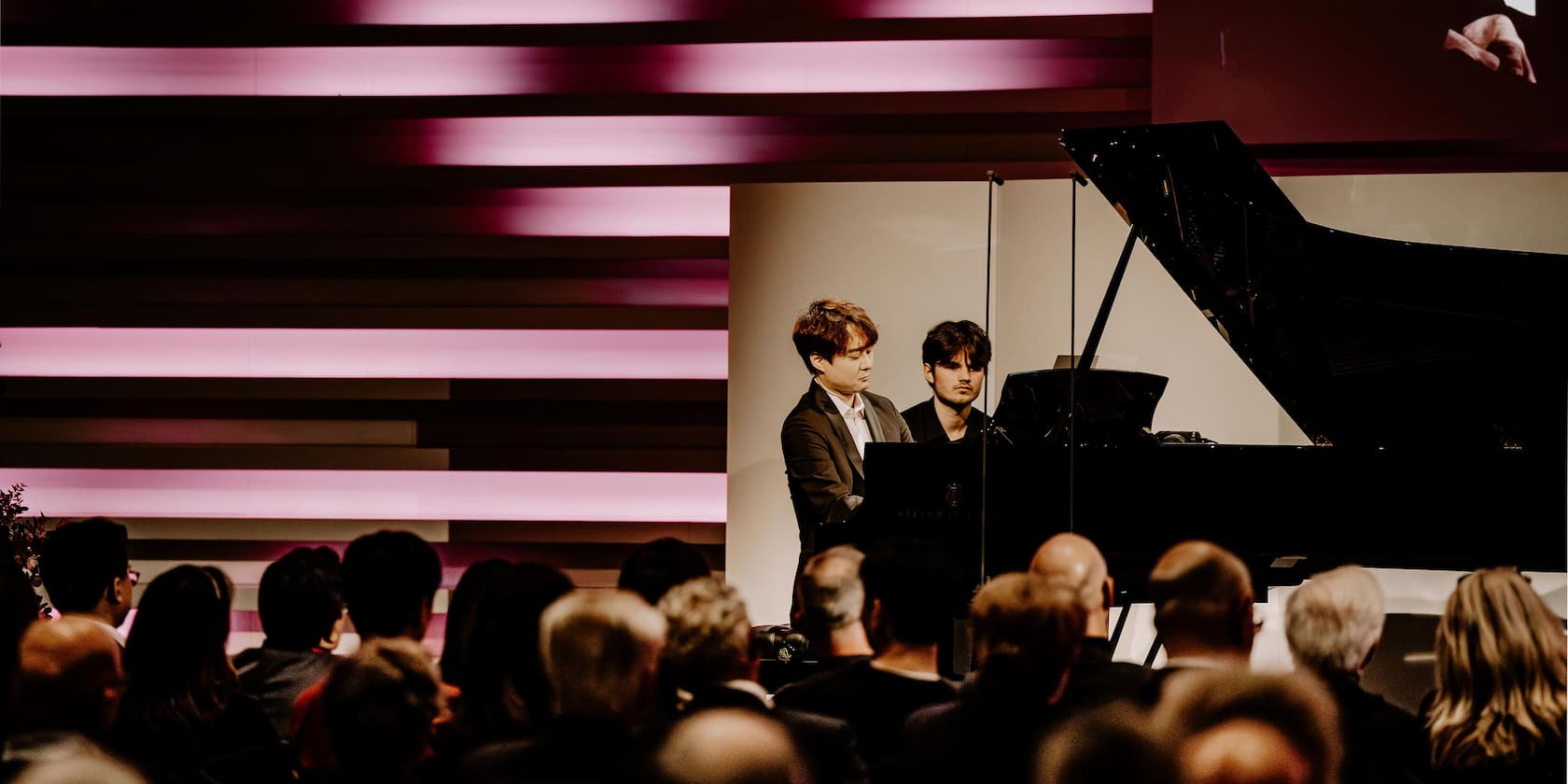 Two pianists play on a stage while an audience listens.