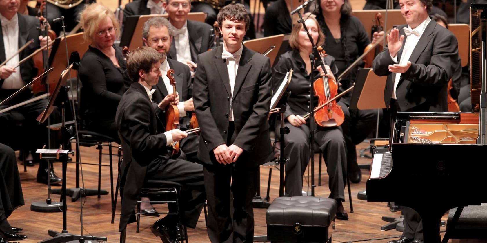 A young man in a suit stands on stage in front of an orchestra and receives applause.