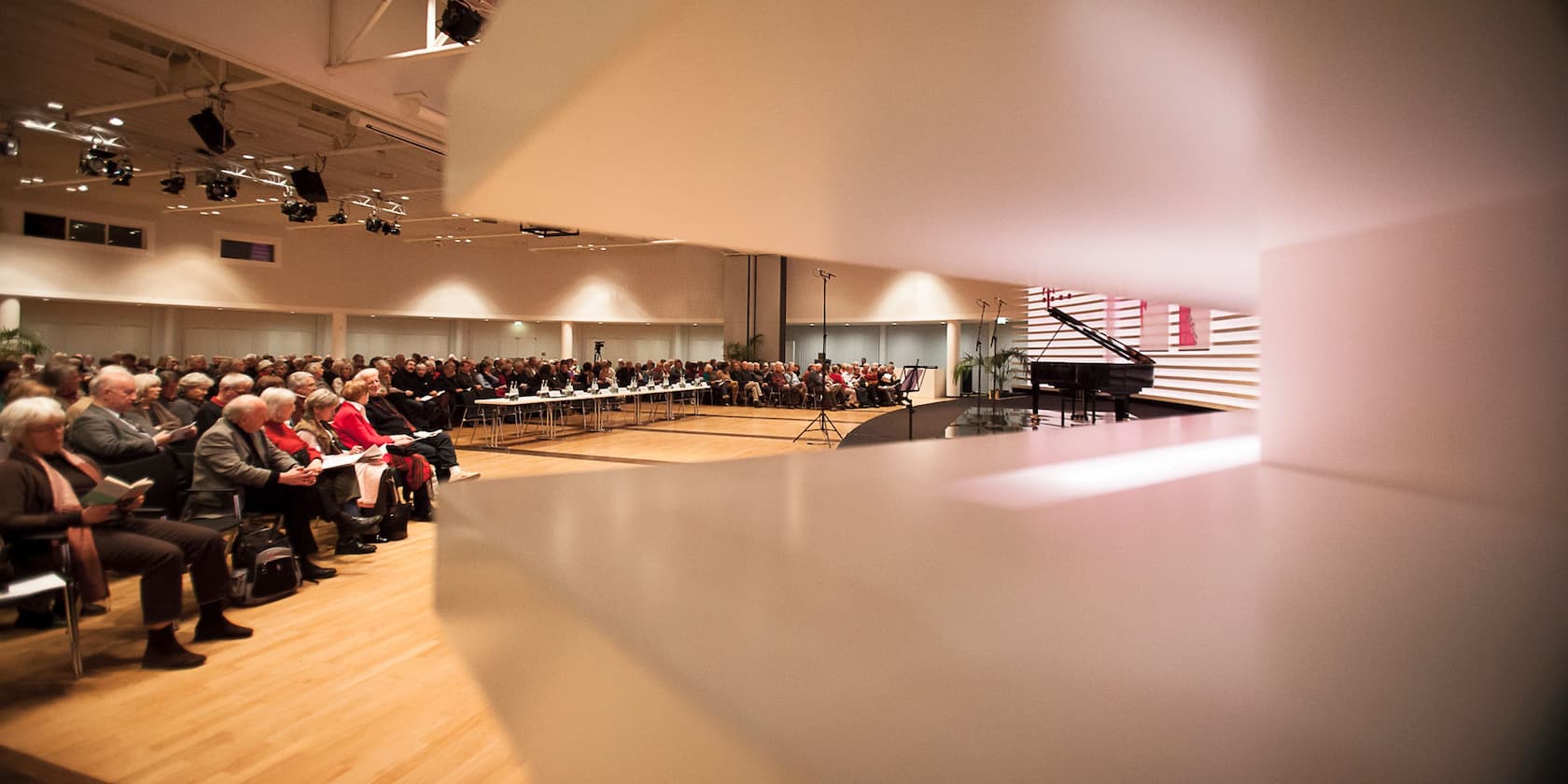 A fully seated event hall with an audience and a grand piano on the stage.