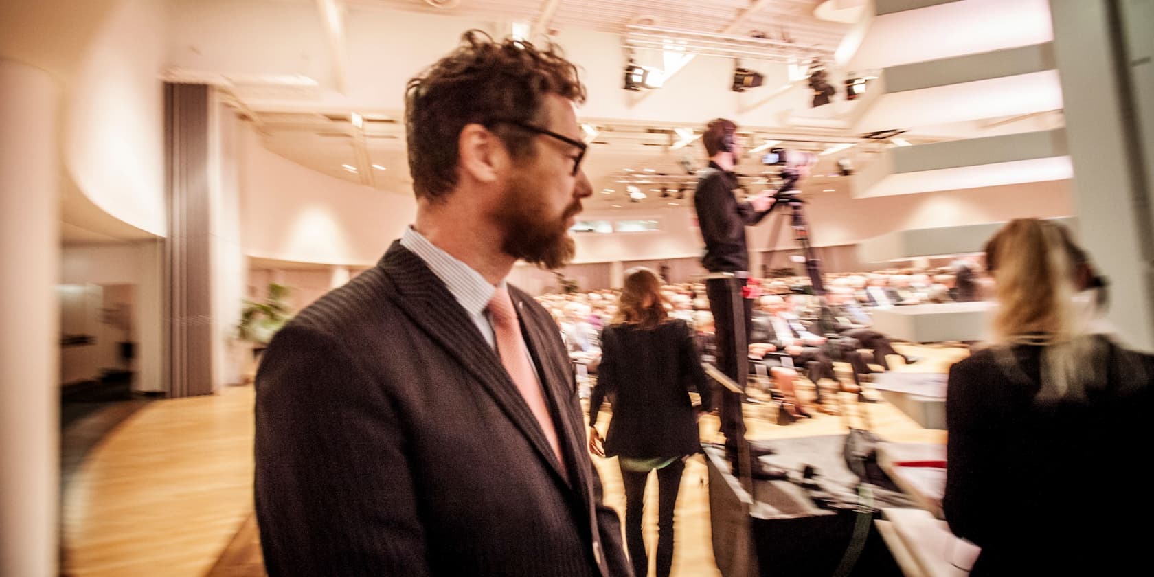 Man in the foreground and a person operating a camera in the background at an event with an audience.