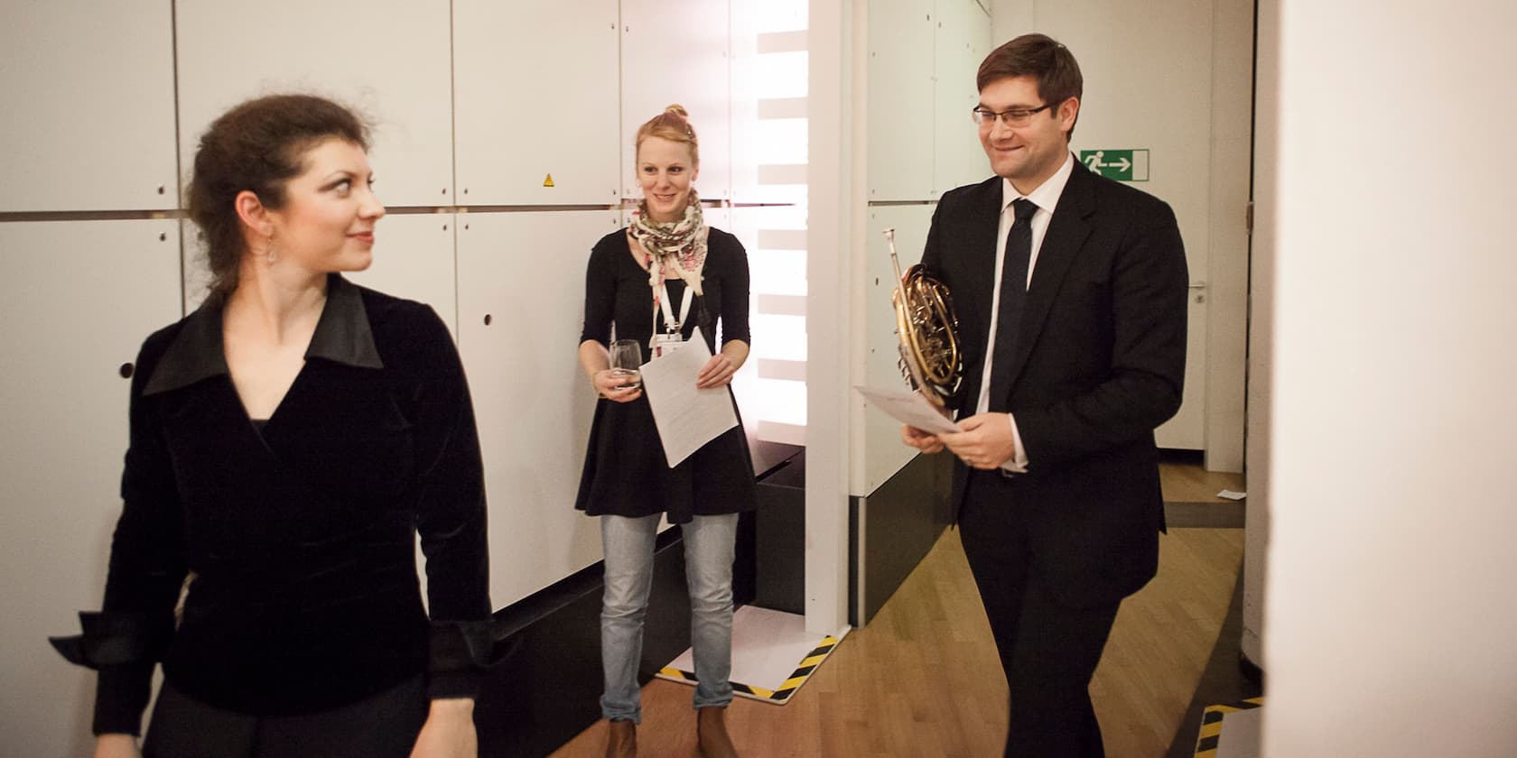 Three people in formal attire conversing in a corridor. One woman holds a glass, and a man holds a horn.