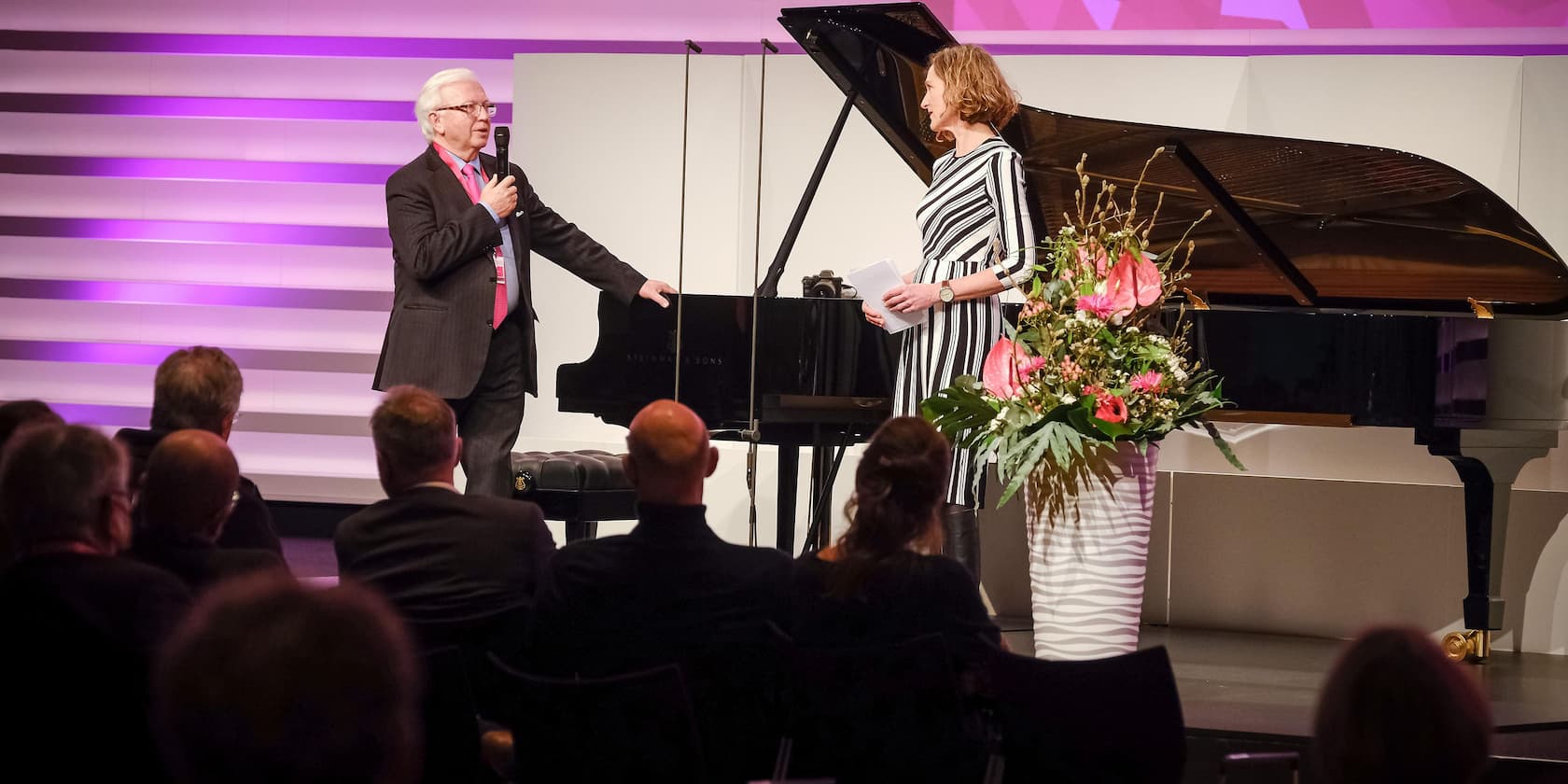 An older man in a suit speaks into a microphone to a woman in a striped dress. In the foreground are a floral arrangement and a grand piano. Audience members are seated in the background.