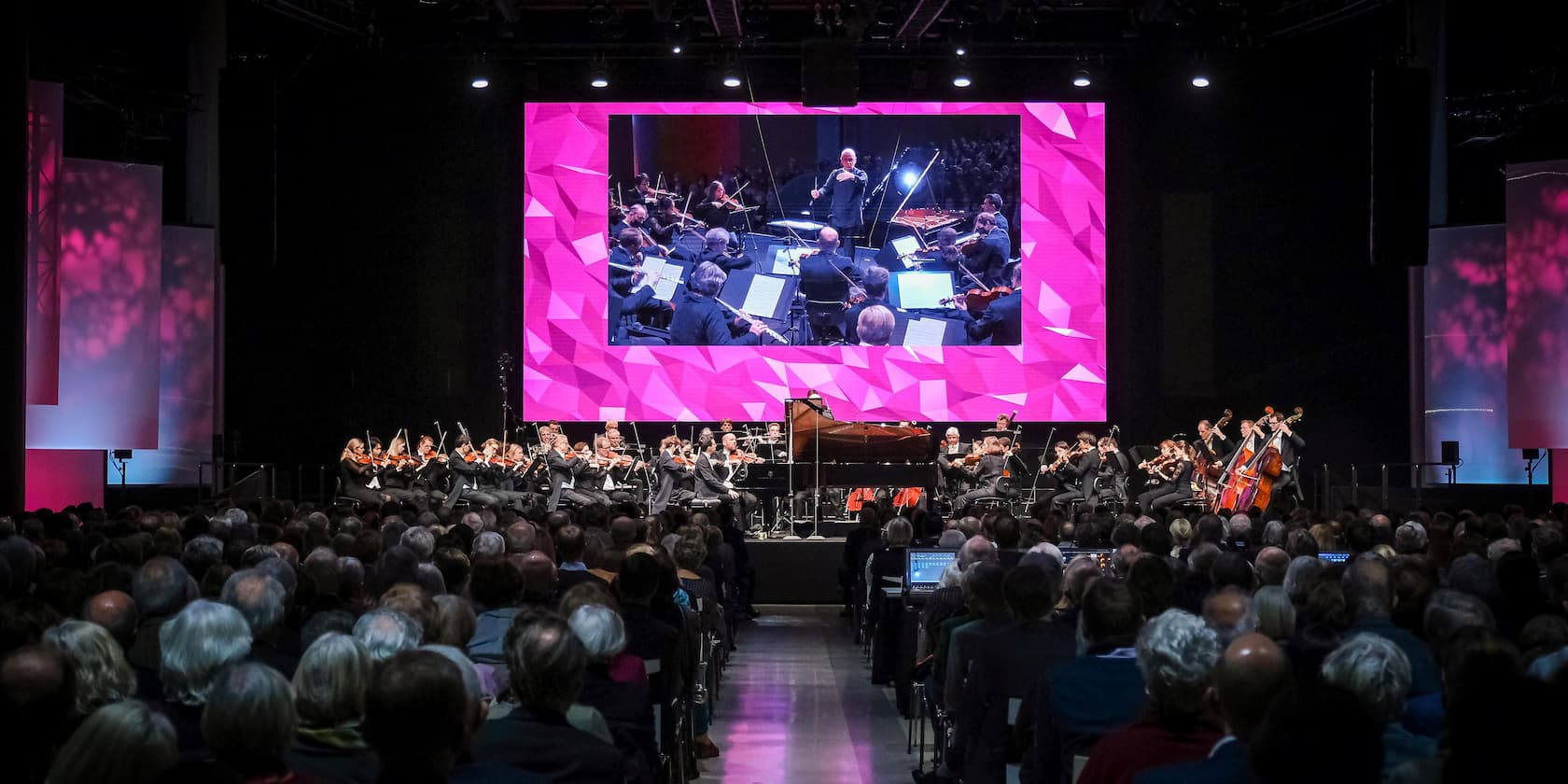 An orchestra performs on stage in front of a large audience. In the background, there is a large screen display of the orchestra with a pink geometric pattern.