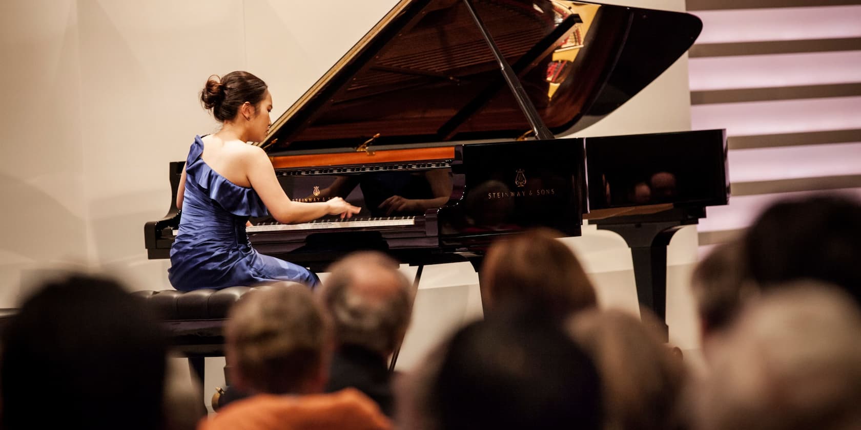 A pianist in a blue dress performing on a grand piano in front of an audience.