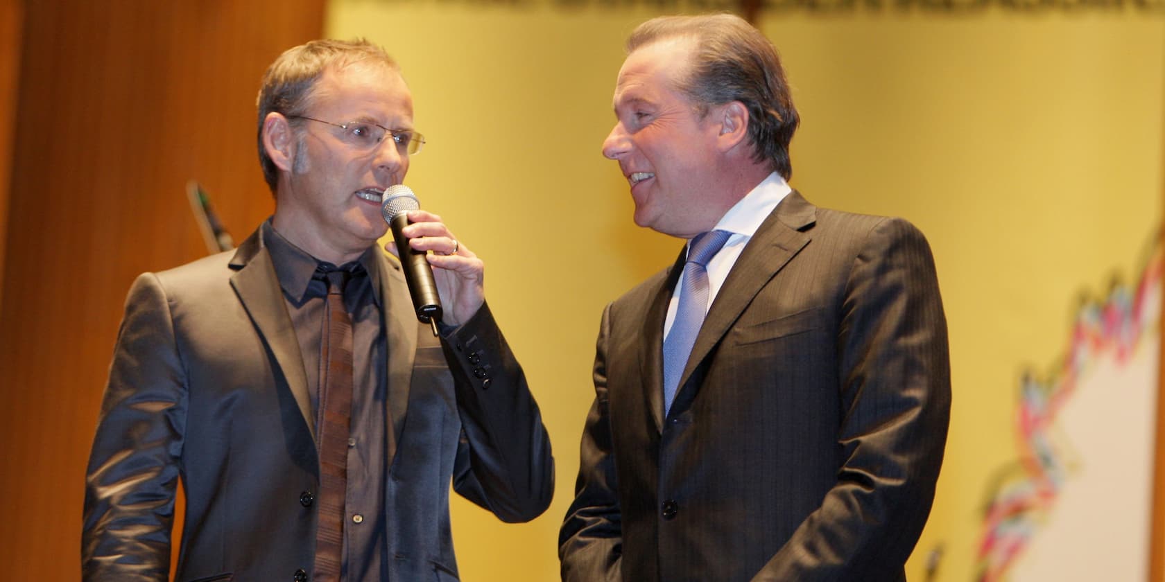 Two men in suits, with one speaking into a microphone, on a stage.