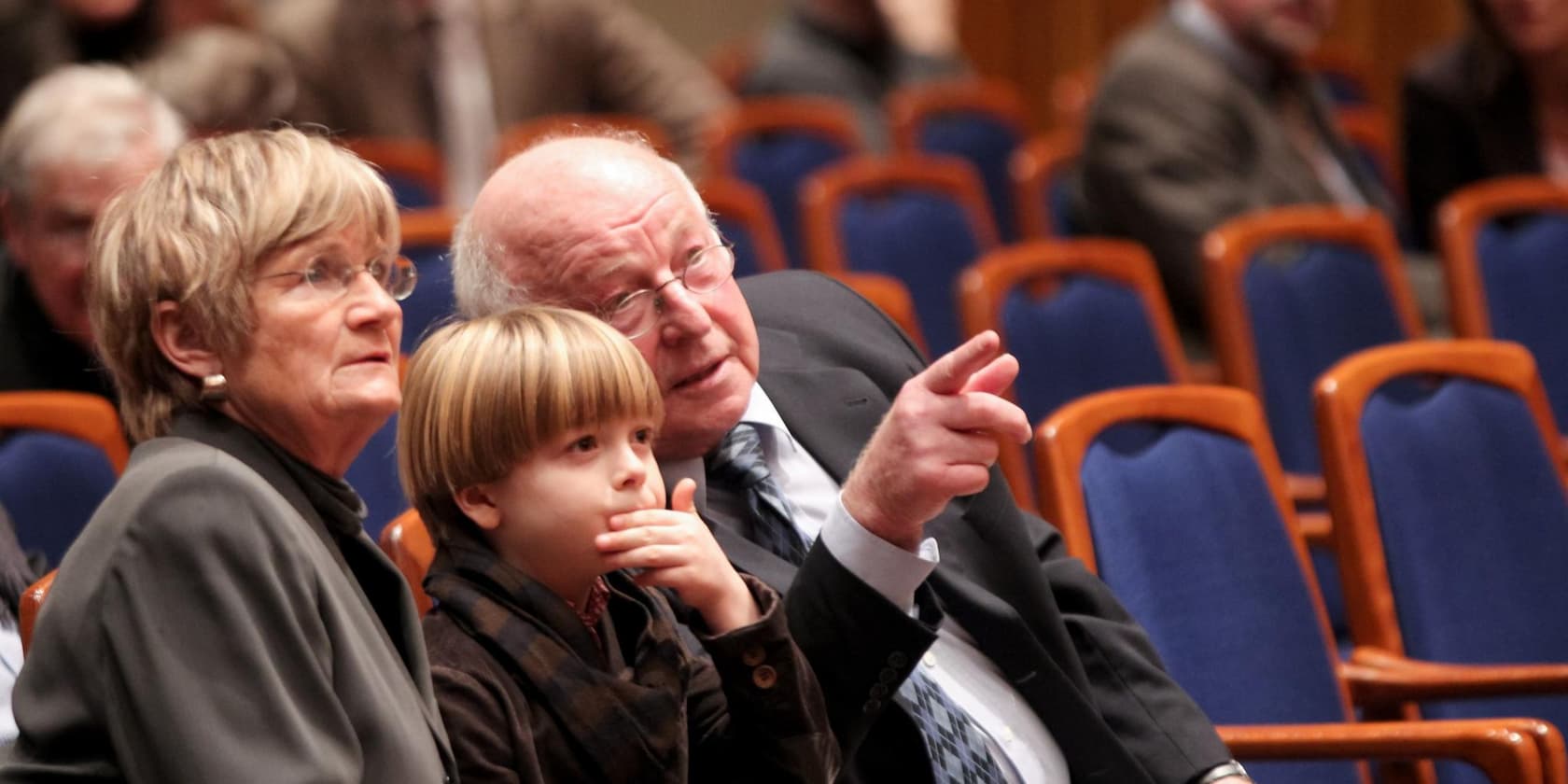 Älteres Paar mit Kind, das in einem Auditorium sitzt. Der ältere Mann zeigt auf etwas.