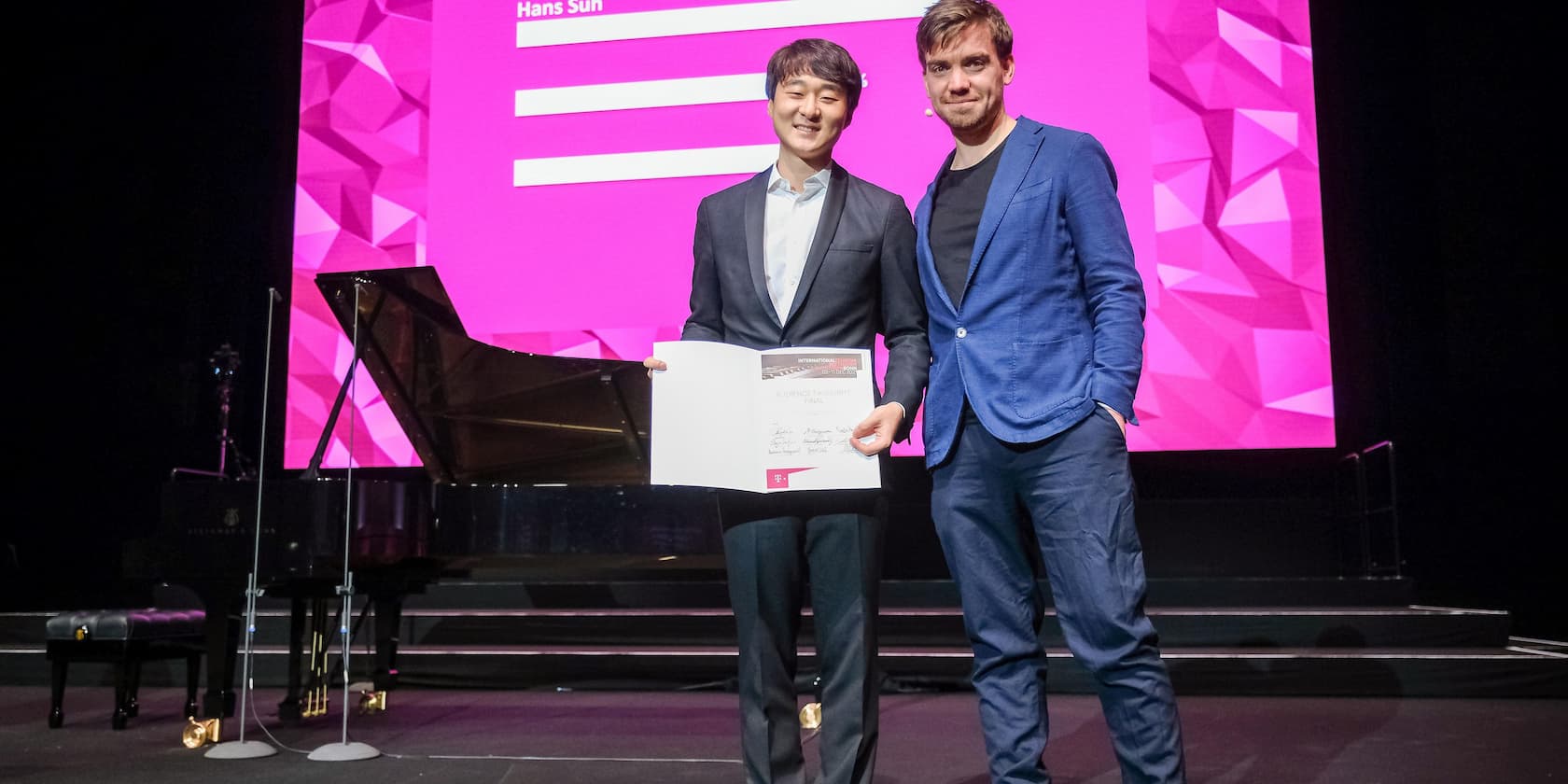 Two men stand on a stage, one holding a certificate. In the background, a piano and a pink screen with a geometric pattern.