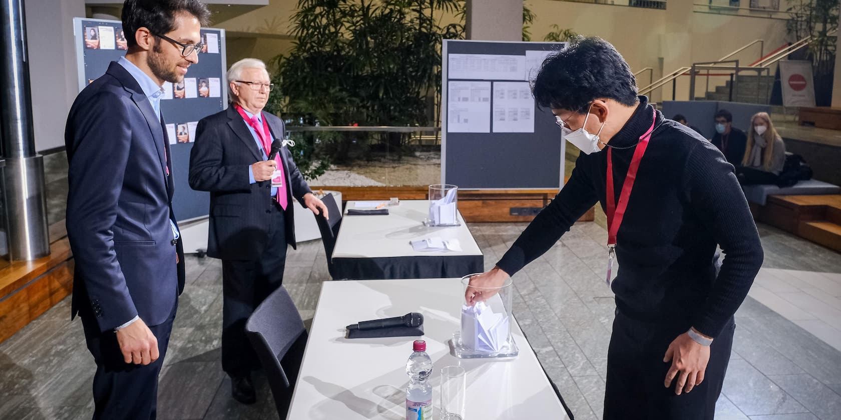 Three men at an event. One of the men inserts a paper into a transparent box while the others watch.