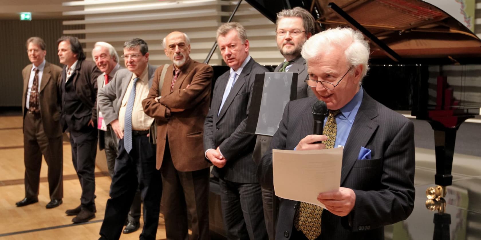 A group of men stands on a stage while one man speaks into a microphone and holds a document.