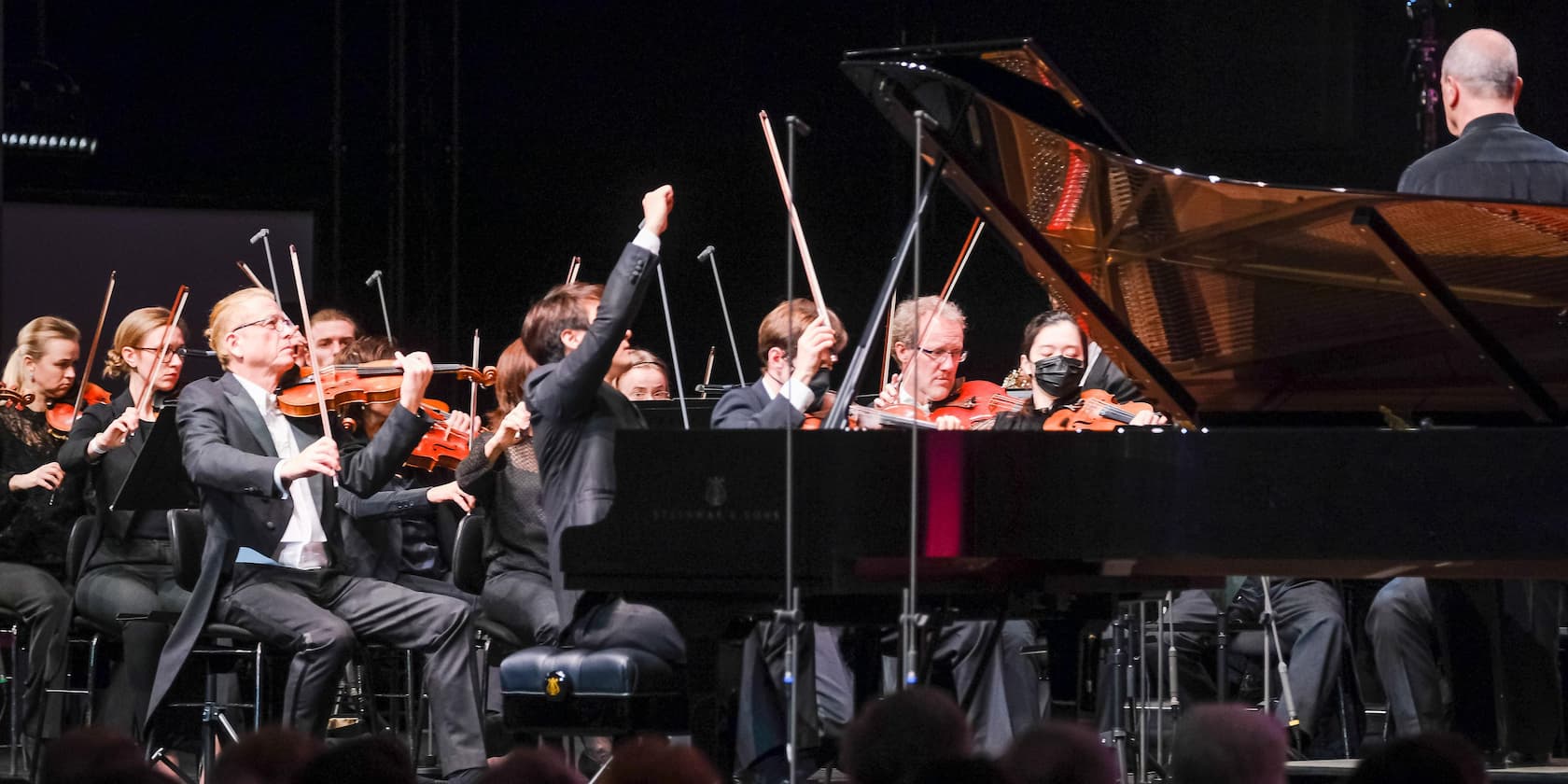 An orchestra is playing on stage while a conductor leads them. A grand piano is visible in the foreground.