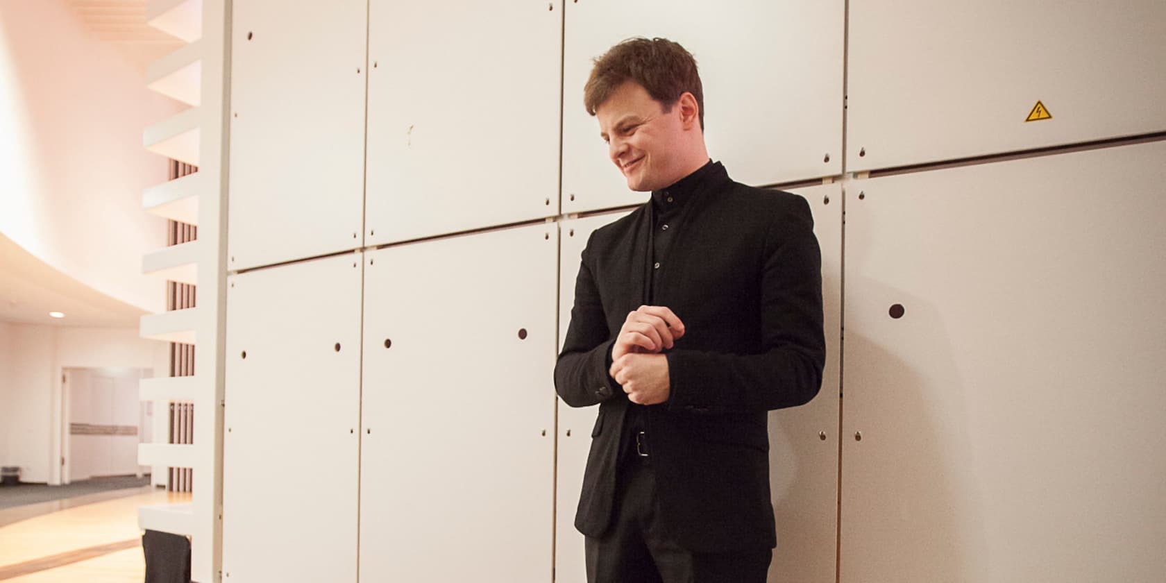 A man in black clothing stands smiling in front of a white wall.