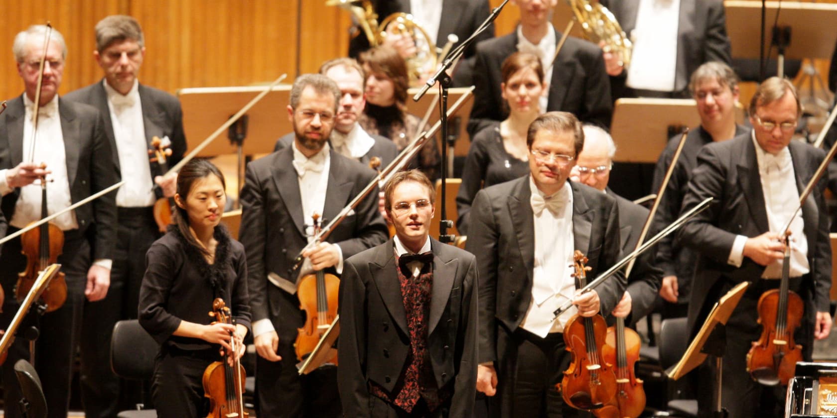 An orchestra stands on a stage, musicians holding their instruments and wearing formal attire.