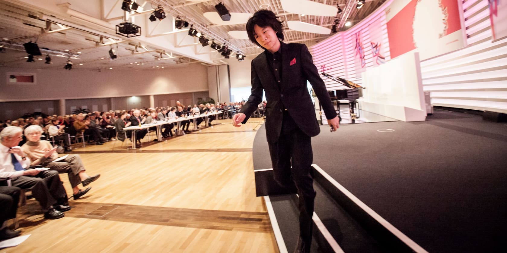 A man in a black suit walks down stairs from a conference stage, while an audience sits at tables.