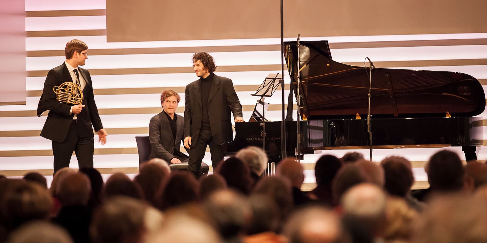 Three musicians on stage in front of an audience. One is holding a horn while two others stand next to a grand piano.