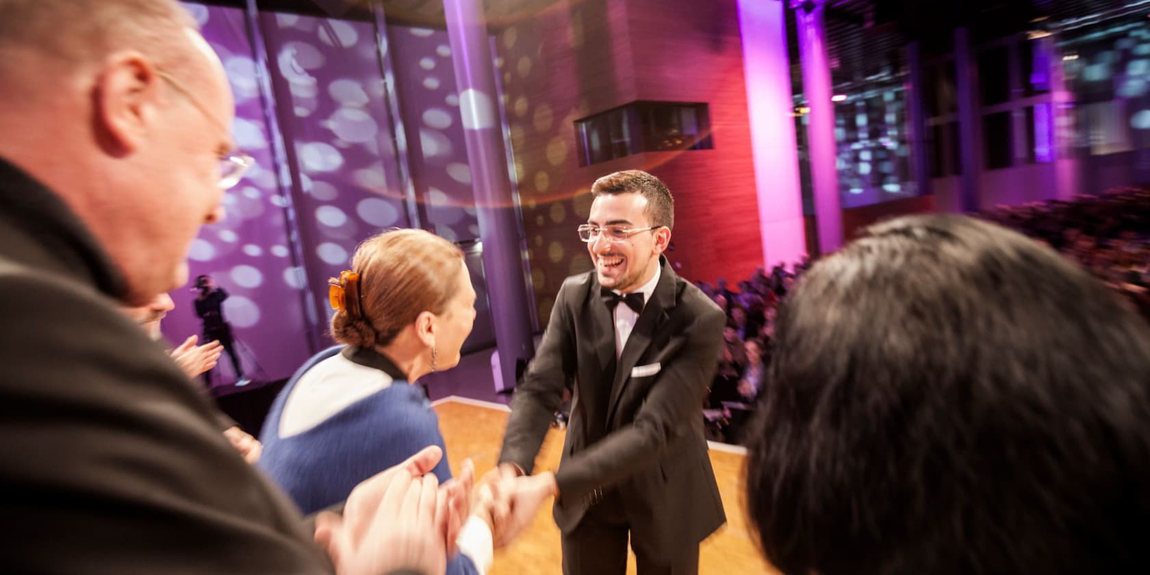 A man in a suit joyfully shakes hands with a woman in front of an audience in a hall with purple lighting.