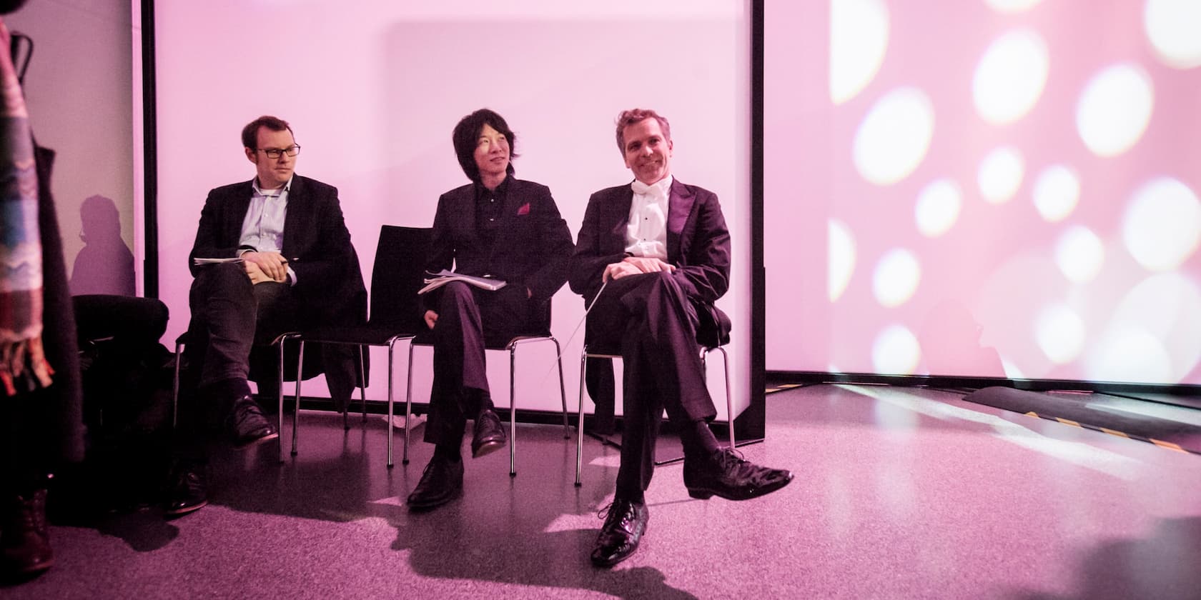 Three men sitting next to each other on chairs against a pink-lit background.