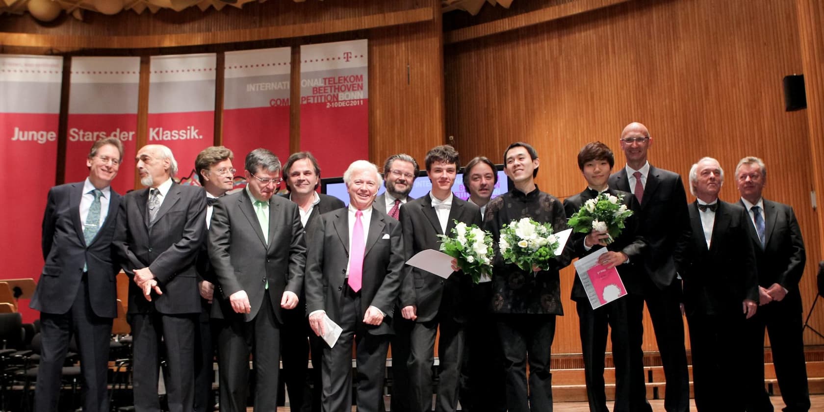 Group photo during a music competition. Two young winners holding bouquets and certificates, surrounded by jury members and organizers. Background banners read: 'Young Stars of Classical Music'.