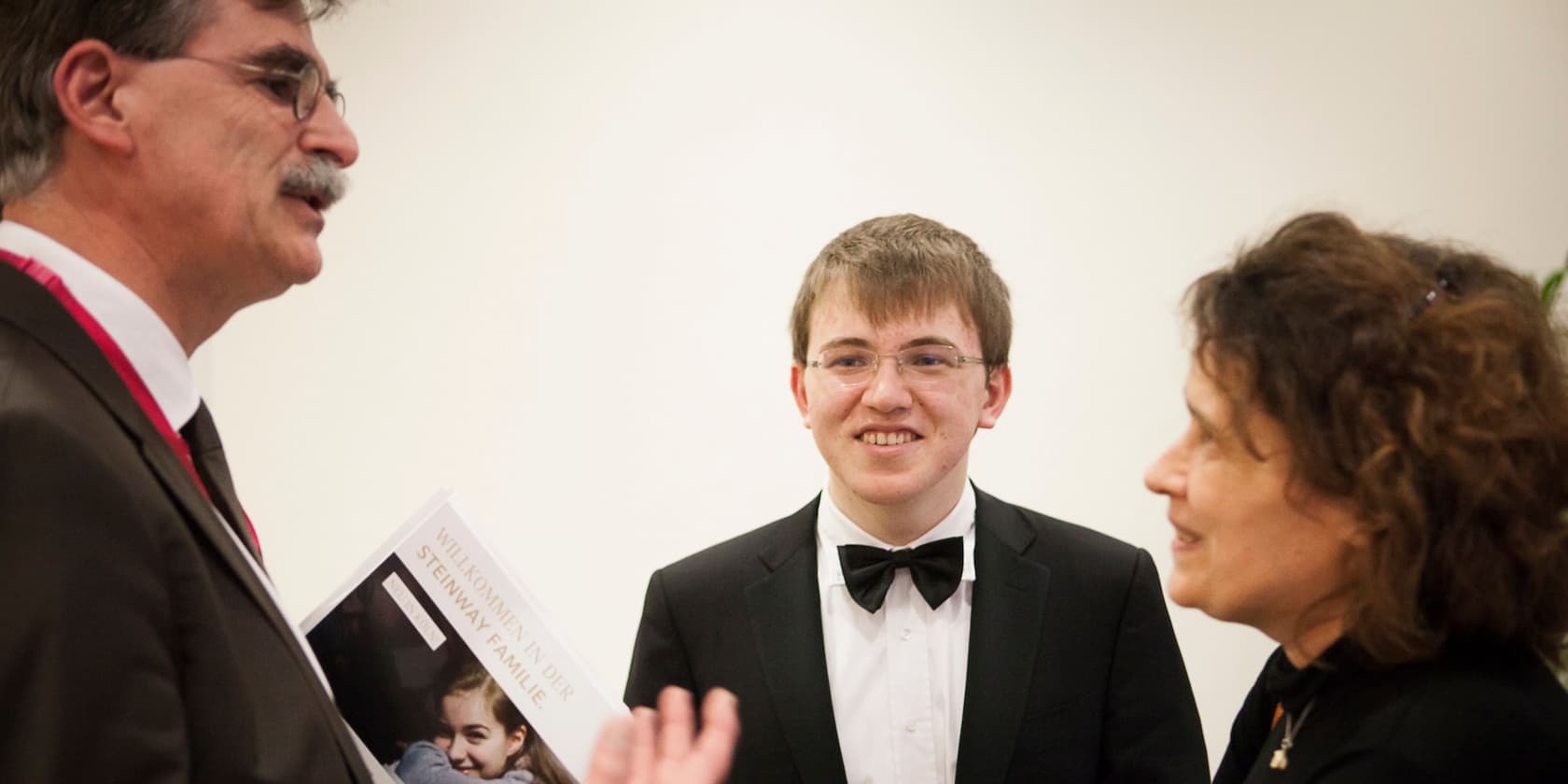 Three people are standing together and talking, one of them is holding a magazine with the title 'Welcome to the Steinway Family'.