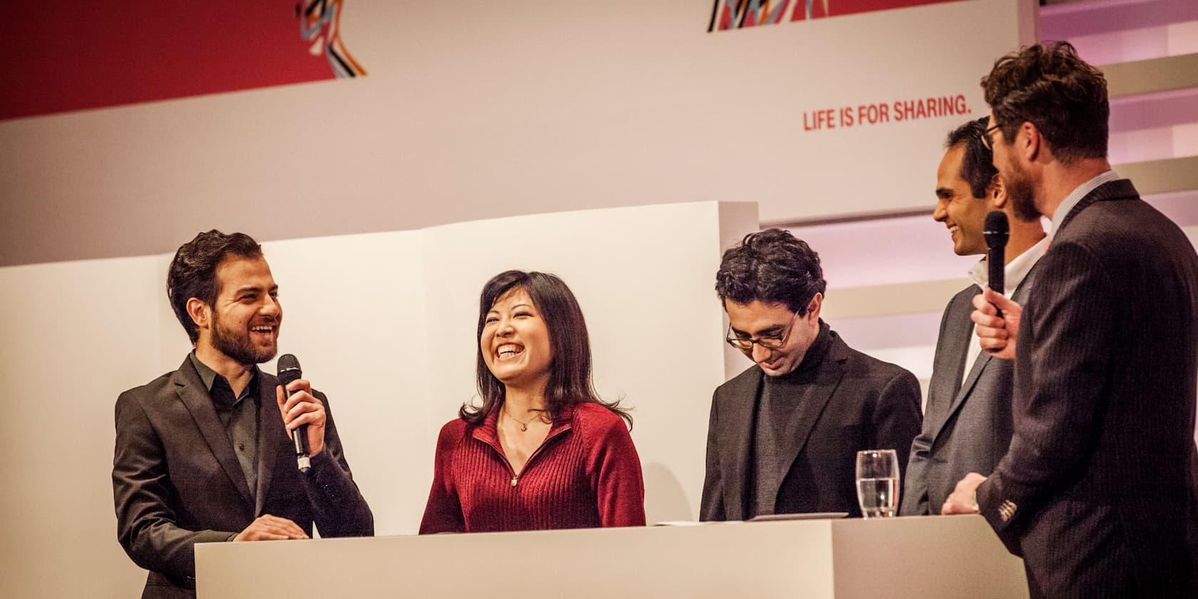 Five people standing and talking on a stage. Two people are holding microphones. The background reads: 'Life is for sharing.'