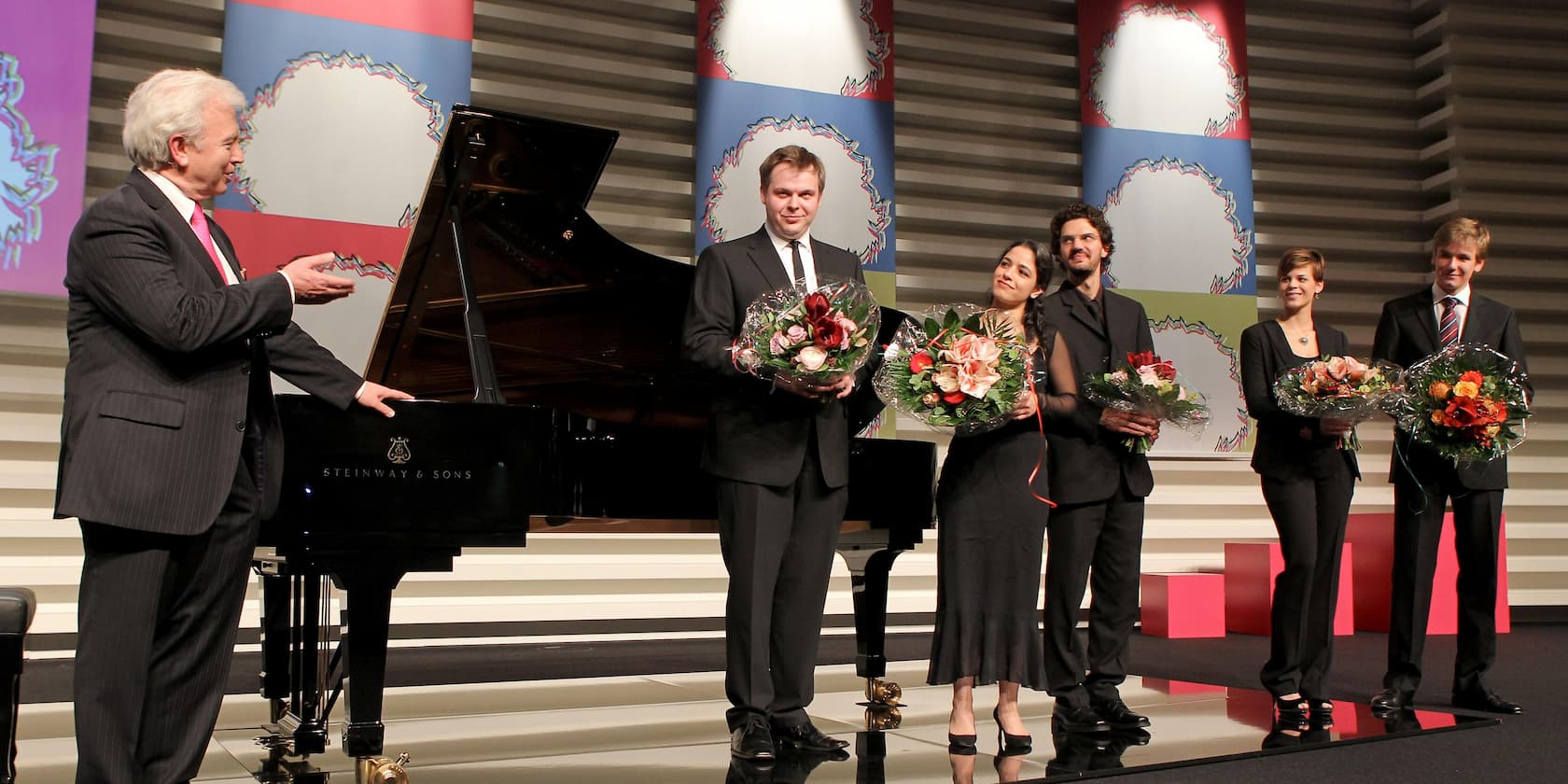 A group of six people at an award ceremony; four individuals are holding bouquets while a man stands next to a Steinway & Sons piano speaking.