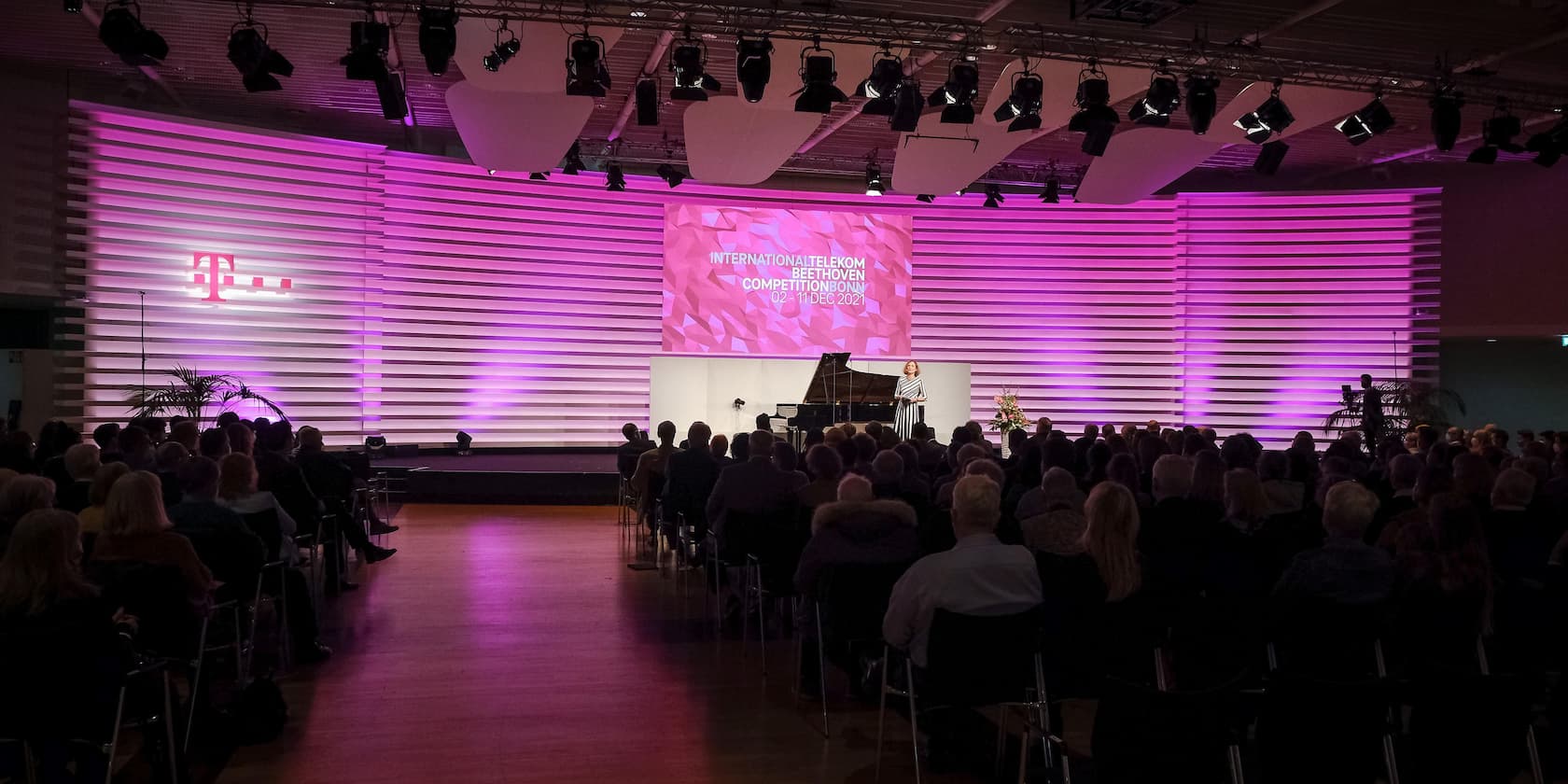 An auditorium with people listening to a woman on stage at the International Telekom Beethoven Competition Bonn. A grand piano and a screen with the text 'INTERNATIONAL TELEKOM BEETHOVEN COMPETITION BONN, 02-11 DEC 2021' are in the background.