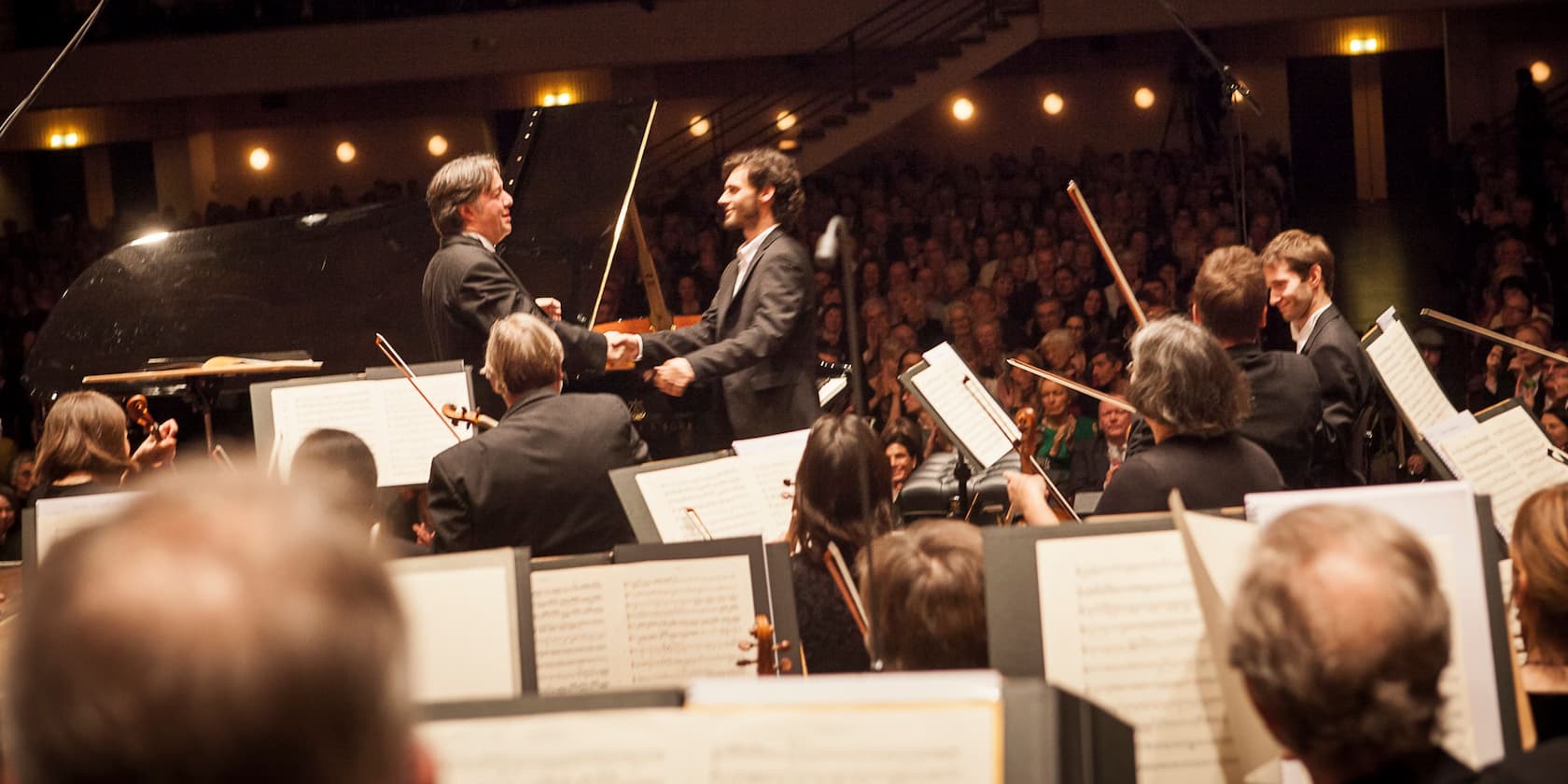 Conductor and pianist shaking hands on stage in front of an orchestra and a large audience.