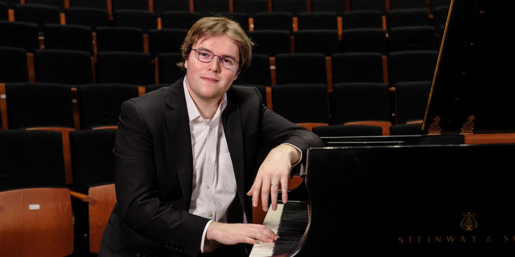 A man sits at a grand piano in a concert hall and looks at the camera.