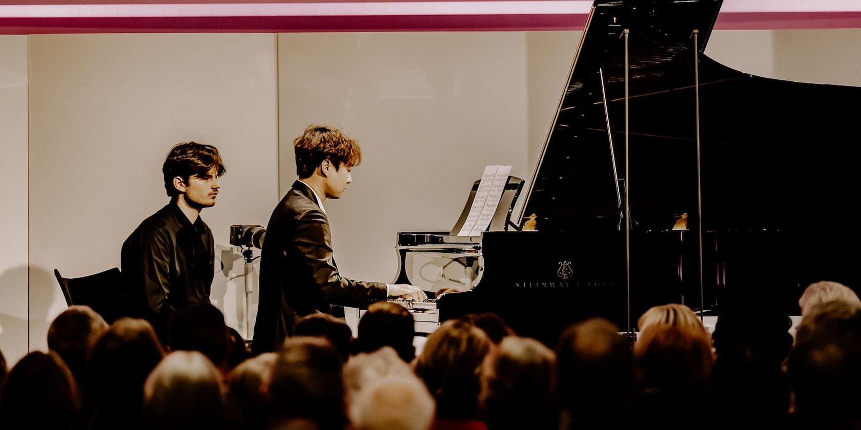 A pianist plays on a grand piano in front of an audience while another person sits beside him.