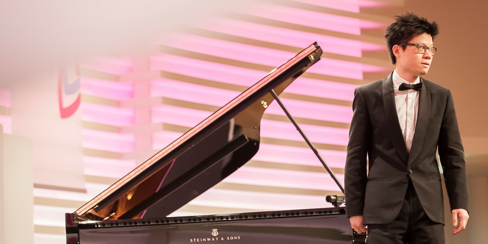 Man in a black suit and bow tie stands next to a grand piano on a stage with pink and white lights.