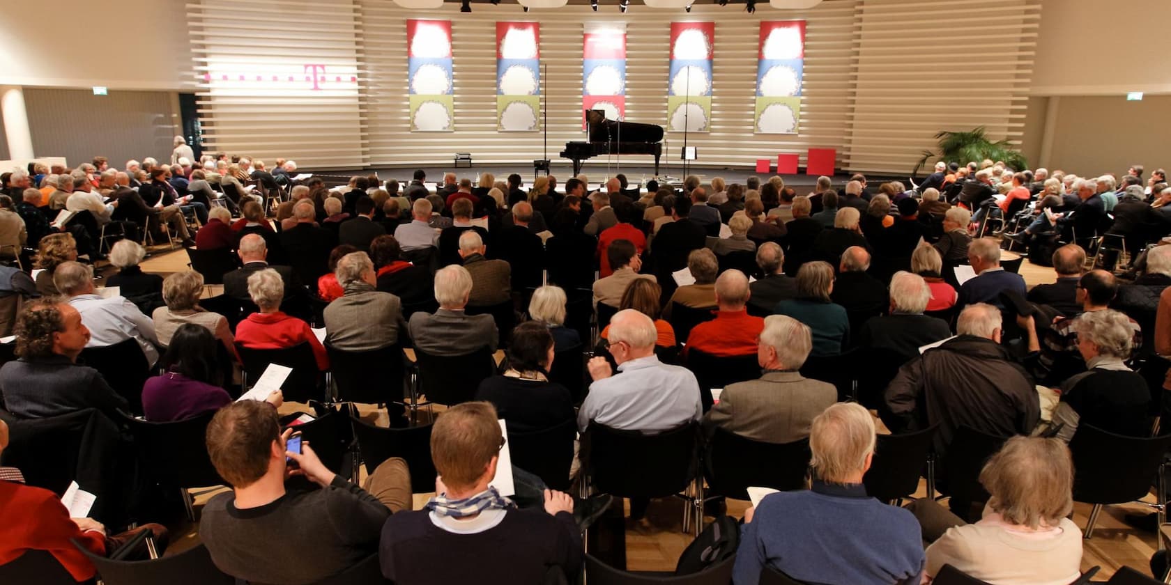 Eine große Gruppe von Menschen sitzt in einem Auditorium und schaut auf eine Bühne mit einem Klavier.