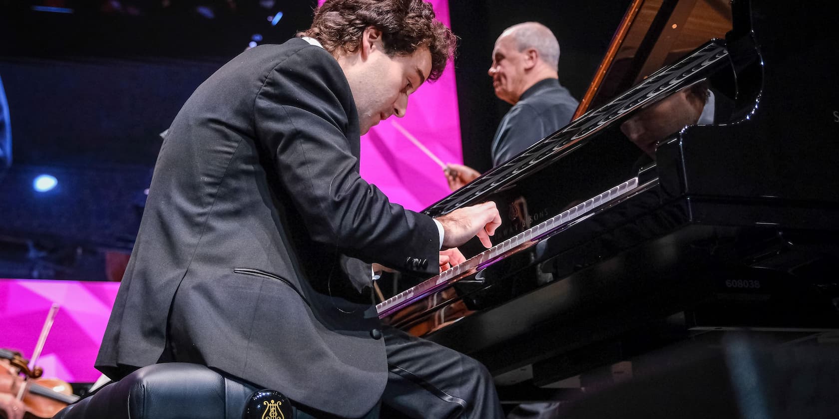 Ein Pianist spielt Klavier auf einer Bühne, mit einem Dirigenten und Orchester im Hintergrund.