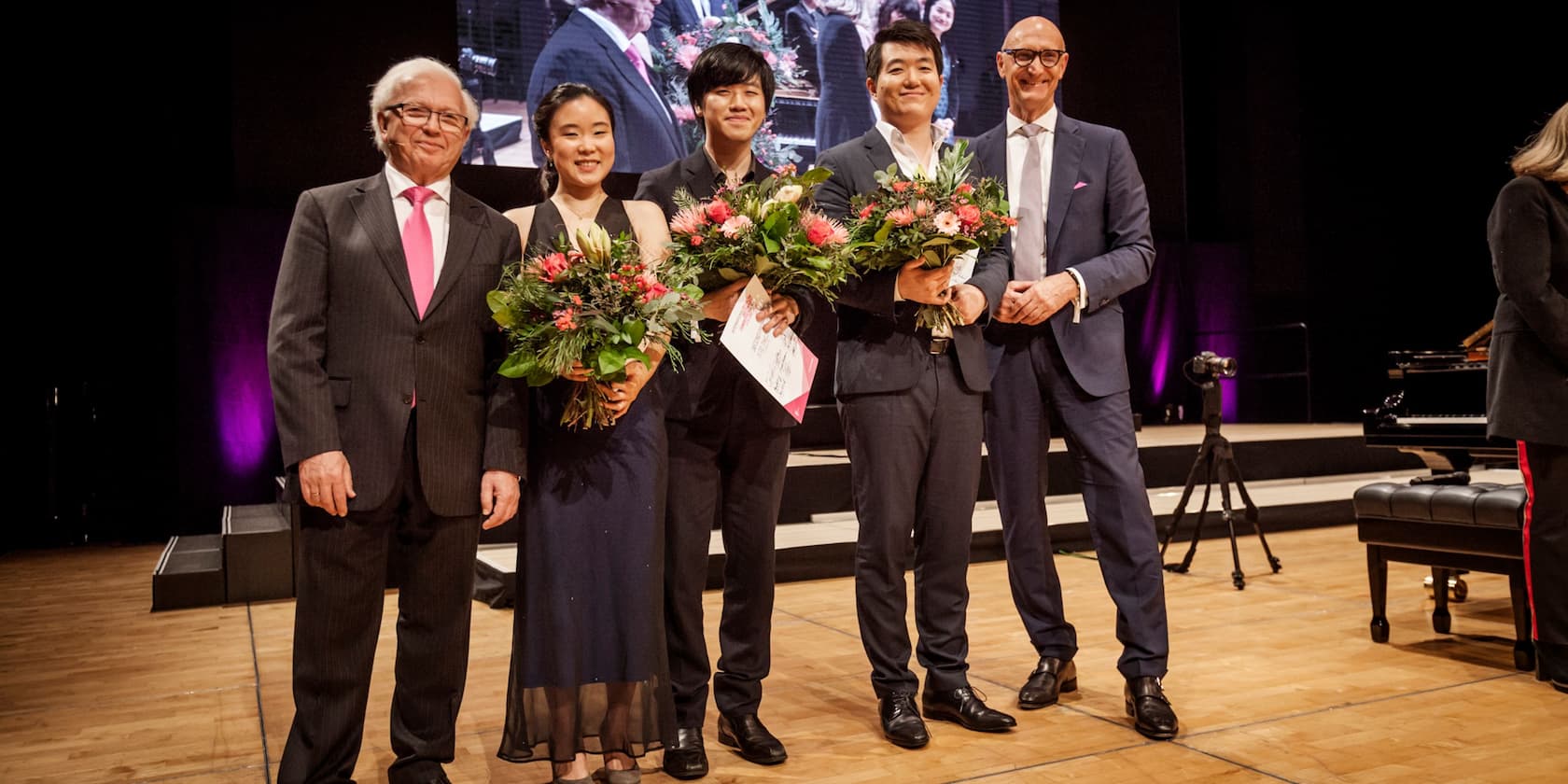 A group of five people standing on a stage, three of them holding bouquets and certificates.