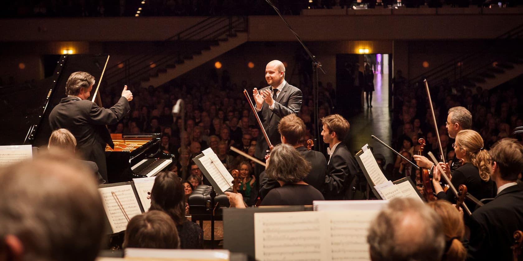 Conductor and pianist are being applauded by the orchestra and audience on a stage in a concert hall.