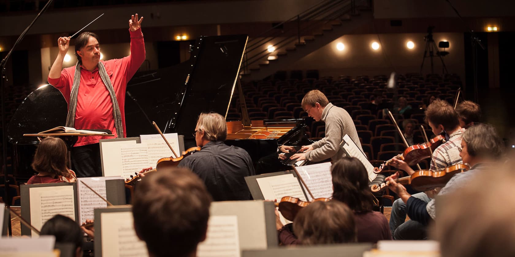 A conductor leads an orchestra during a rehearsal. A pianist plays on a grand piano.