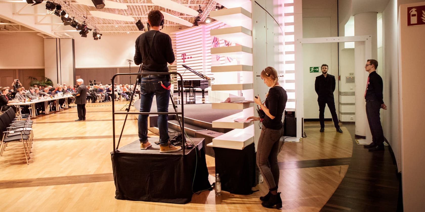 Preparations for a conference in a large hall. A person is filming the event while participants sit at tables and listen to a speaker.