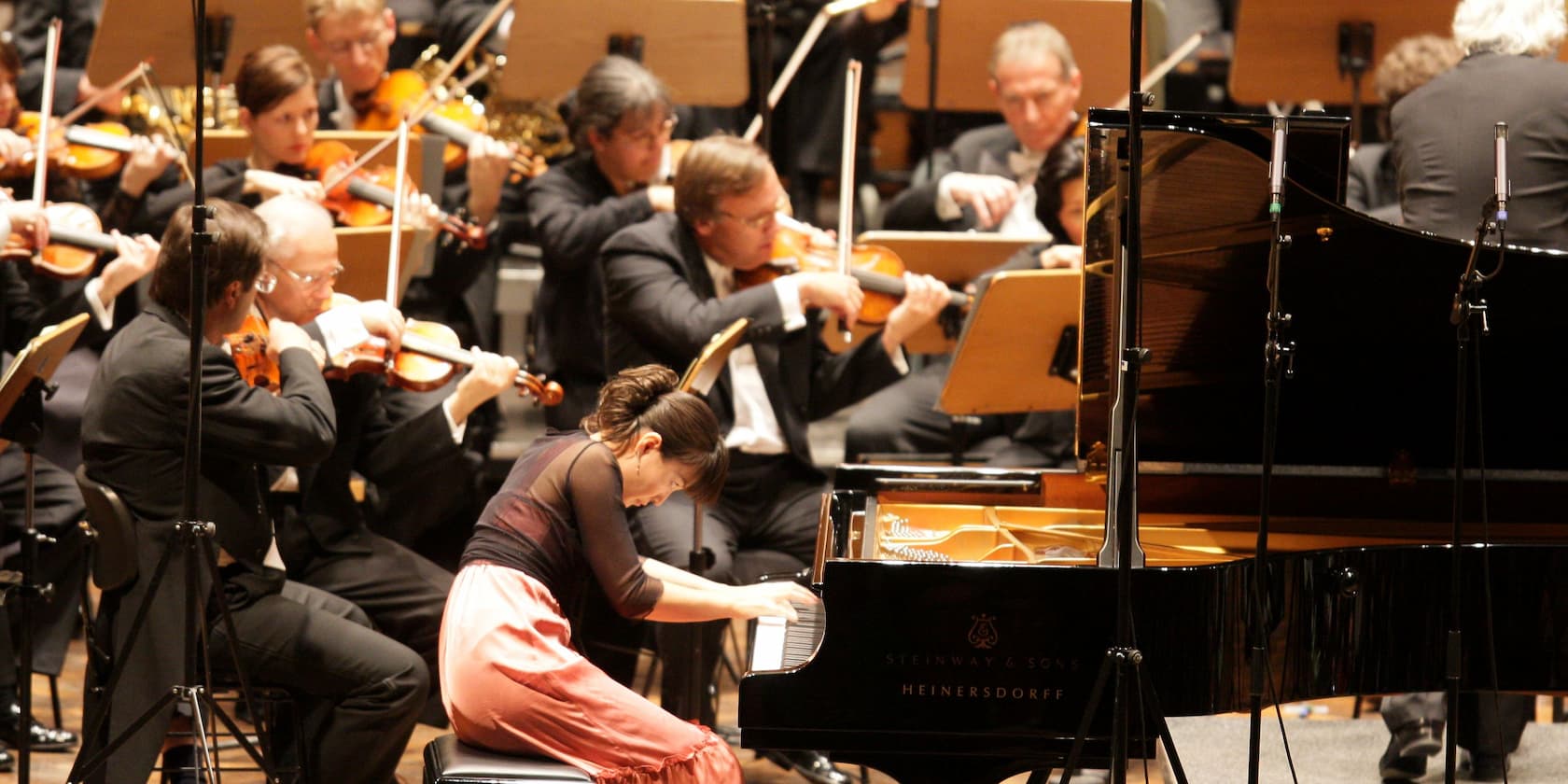 A woman plays the piano on stage, surrounded by an orchestra with violinists.