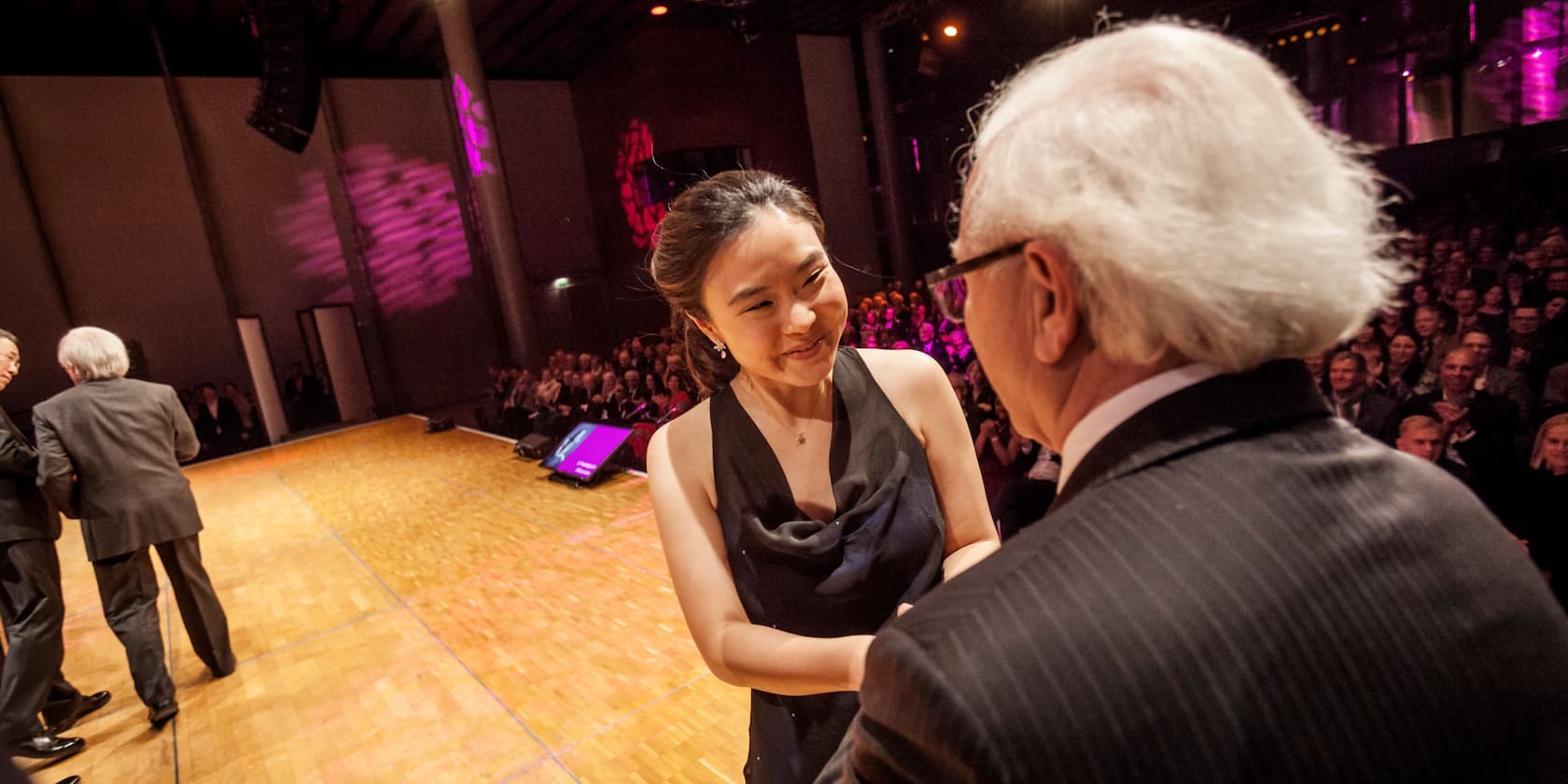 A woman in a black dress is greeted by an older man on a stage while the audience applauds.